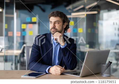 A thoughtful businessman in a modern office setting, contemplating strategy. He is surrounded by digital devices and colorful notes on a glass wall, reflecting a dynamic work atmosphere. 118175651