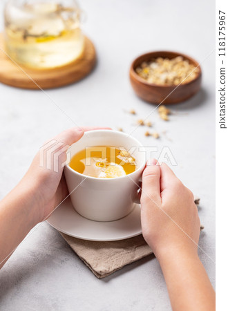 Hands holding a glass cup with chamomile herbal tea on a light background with dry flowers  118175967