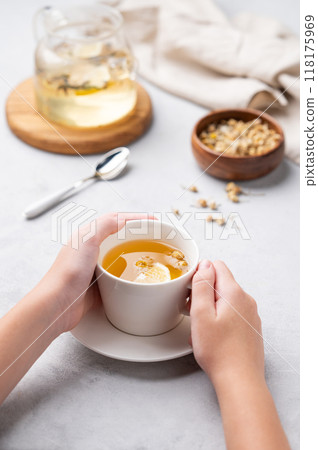 Hands holding a glass cup with chamomile herbal tea on a light background with dry flowers Hands holding a glass cup with chamomile herbal tea on a light background with dry flowers 118175969
