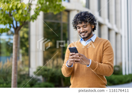 Young professional with curly hair using smartphone outdoors. Scene conveys connectivity, technology, and communication. Individual dressed in casual autumn attire, standing confidently near office 118176411