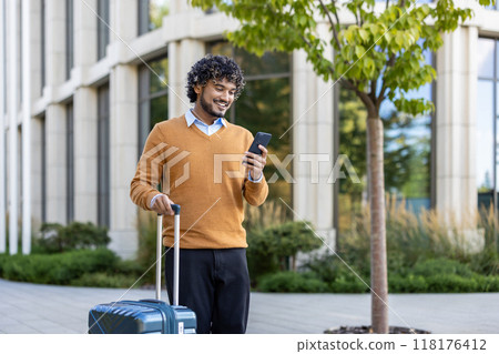 Smiling man checks phone while pulling suitcase outside modern building. Represents business travel, technology use, and professional lifestyle in casual attire, connecting work and leisure Smiling man checks phone while pulling suitcase outside modern building. Represents business travel, technology use, and professional lifestyle in casual attire, connecting work and leisure 118176412