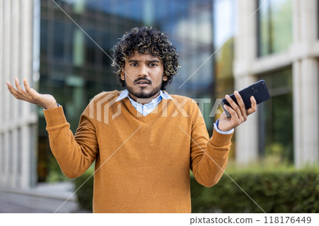 Man with curly hair in sweater holding smartphone with confused expression. Outdoor urban background suggests business context, conveying uncertainty, doubt, or perplexity in modern communication. Man with curly hair in sweater holding smartphone with confused expression. Outdoor urban background suggests business context, conveying uncertainty, doubt, or perplexity in modern communication. 118176449