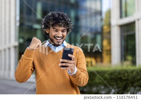 Happy young man displaying excitement while looking at smartphone in city environment. Man wearing sweater celebrates success or good news. Captures moment of digital connection and achievement. Happy young man displaying excitement while looking at smartphone in city environment. Man wearing sweater celebrates success or good news. Captures moment of digital connection and achievement. 118176451