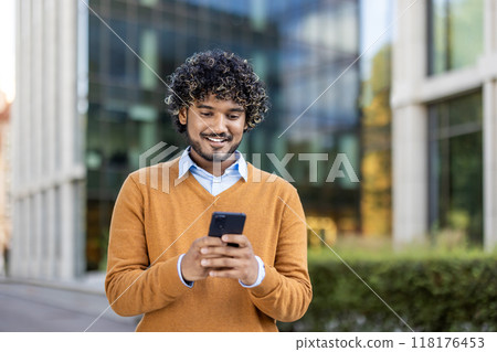 Young professional smiling while using smartphone in urban setting. Dressed in casual attire, enjoying communication and connectivity during outdoor break near office buildings. 118176453