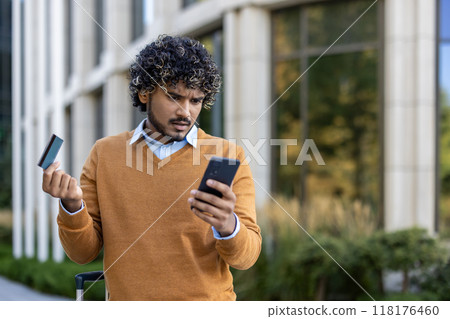 Man with curly hair wearing brown sweater holding credit card and smartphone. Standing outdoors appearing confused, possibly facing online banking or payment problem during business trip. 118176460