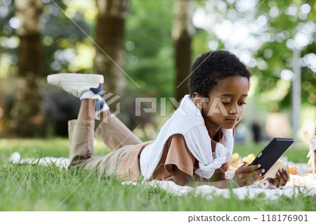 Little African American boy using smartphone and eating sandwich while lying on blanket in park 118176901