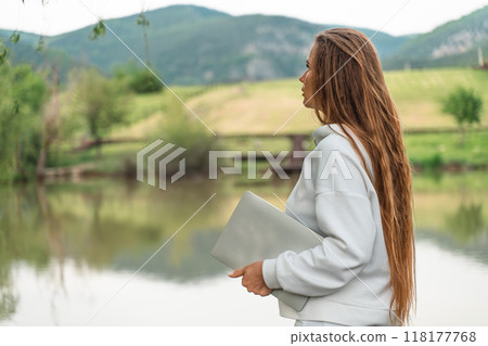 A woman with long hair is standing by a lake, holding a laptop. Concept of solitude and contemplation, as the woman is lost in thought while gazing out at the water. A woman with long hair is standing by a lake, holding a laptop. Concept of solitude and contemplation, as the woman is lost in thought while gazing out at the water. 118177768