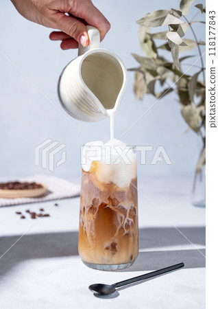 Iced coffee latte in a tall glass with a hand pouring milk from a milk jug on a blue background 118177843