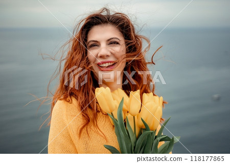 Portrait of a happy woman with hair flying in the wind against the backdrop of mountains and sea. Holding a bouquet of yellow tulips in her hands, wearing a yellow sweater Portrait of a happy woman with hair flying in the wind against the backdrop of mountains and sea. Holding a bouquet of yellow tulips in her hands, wearing a yellow sweater 118177865