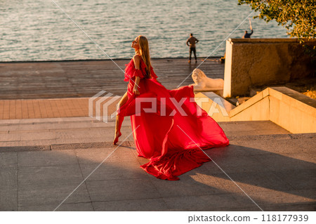 Sunrise red dress. A woman in a long red dress against the backdrop of sunrise, bright golden light of the sun's rays. The concept of femininity, harmony. 118177939