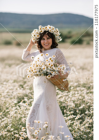 Happy woman in a field of daisies with a wreath of wildflowers on her head. woman in a white dress in a field of white flowers. Charming woman with a bouquet of daisies, tender summer photo Happy woman in a field of daisies with a wreath of wildflowers on her head. woman in a white dress in a field of white flowers. Charming woman with a bouquet of daisies, tender summer photo 118177975