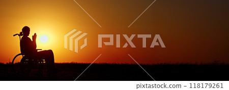 Silhouette of a person in a wheelchair at sunset with hands clasped, set against a vivid orange sky. 118179261