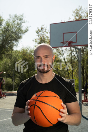 Portrait of sporty man in black t-shirt holding basketball orange ball and looking at camera on sports court. Basketball coach. Close up. Outdoors. 118179557