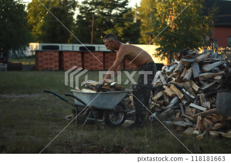 A man is seen working with a wheelbarrow filled with firewood, assembling and stacking a large pile. he organizes the stack of wood A man is seen working with a wheelbarrow filled with firewood, assembling and stacking a large pile. he organizes the stack of wood 118181663