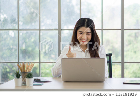 Young asian woman in white shirt sitting at a desk with laptop, smiling and raise finger thumbs up at the screen, engaged in a video call or virtual meeting. Young asian woman in white shirt sitting at a desk with laptop, smiling and raise finger thumbs up at the screen, engaged in a video call or virtual meeting. 118181929