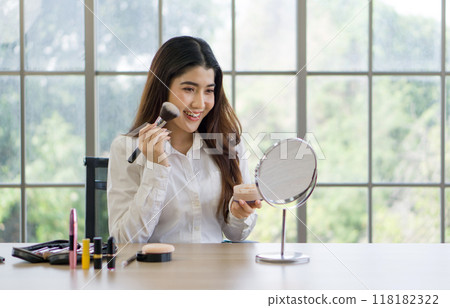 A woman is happily applying makeup while looking in a mirror at a table with various beauty products. 118182322