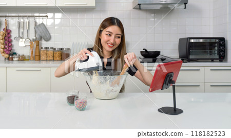 Young asian woman with a smile baking in a kitchen, using an electric mixer and a mixing bowl while looking at online cooking tutorial vdo on tablet computer. 118182523