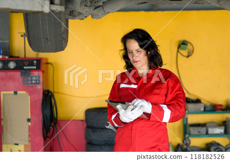 A woman car mechanic stand beneath a lifted car in a garage, typing on tablet computer surrounded by tools A woman car mechanic stand beneath a lifted car in a garage, typing on tablet computer surrounded by tools 118182526