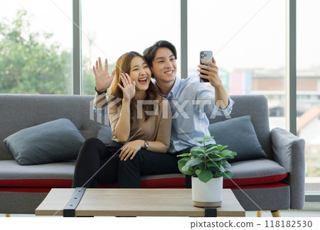Young asian couple sitting on a couch, waving hand via remote communication through smartphone, in a bright living room. Young asian couple sitting on a couch, waving hand via remote communication through smartphone, in a bright living room. 118182530