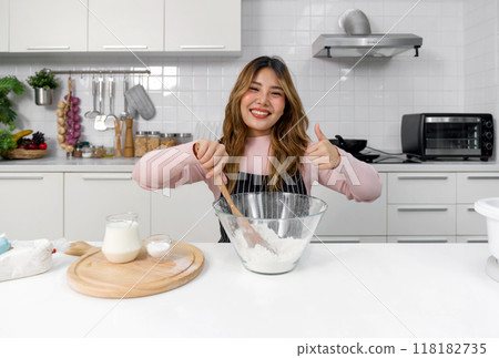 A happy woman in an apron, mixing batter in a clear glass bowl with spatula. Sitting in a clean and modern kitchen, smile while raise finger thumb up. Tray of milk and baking powder placed beside. 118182735