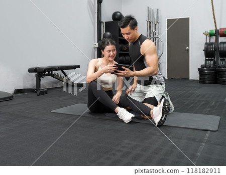 Young asian female sitting on the rubber floor in the gym, check the exercise schedule through tablet computer with male instructor. 118182911