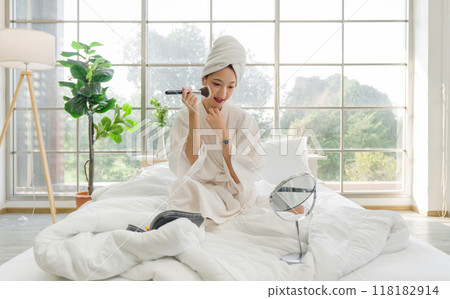 Young asian woman sit relax on a bed in white robe and towel wrapped around her head, doing makeup with a mirror and makeup zippered case. Young asian woman sit relax on a bed in white robe and towel wrapped around her head, doing makeup with a mirror and makeup zippered case. 118182914