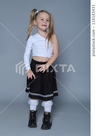A young girl with blonde hair styled in pigtails wears a trendy white one-sleeve top and a black pleated skirt. Her playful stance and subtle smile capture the joy of youth. 118183431