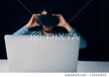 Blurred man in VR headset operating laptop in dark setting, depicting virtual reality technology use. 118183633