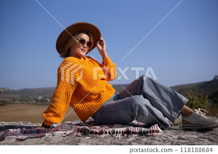 A woman in a yellow sweater and blue jeans is sitting on a blanket on a hillside 118188684