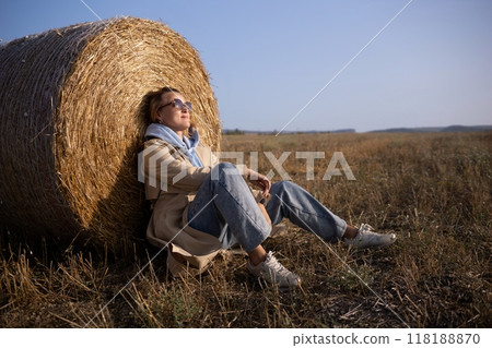 A woman is sitting on a hay bale in a field A woman is sitting on a hay bale in a field 118188870