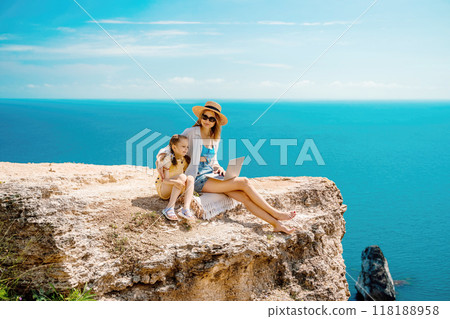 A woman and a child are sitting on a rock overlooking the ocean. The woman is using a laptop while the child looks on. Concept of relaxation and bonding between the two. A woman and a child are sitting on a rock overlooking the ocean. The woman is using a laptop while the child looks on. Concept of relaxation and bonding between the two. 118188958