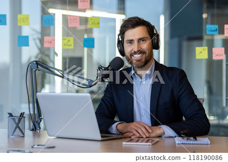 Smiling male podcaster in office setting recording business podcast. Wearing headphones, speaking into microphone. Suitable for themes of communication, entrepreneurship, and media production. Smiling male podcaster in office setting recording business podcast. Wearing headphones, speaking into microphone. Suitable for themes of communication, entrepreneurship, and media production. 118190086
