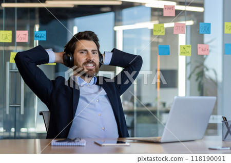 Business professional enjoying a moment of relaxation in modern office setting. Headphones on, laptop open, sticky notes on glass wall symbolize productivity and creativity. 118190091
