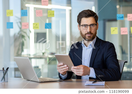 Businessman in suit holding tablet in hand, working with laptop in modern office. Notes on glass wall suggest collaborative work environment. 118190099