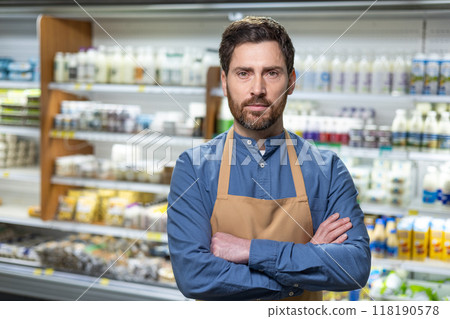 Grocery store employee stands confidently in front of dairy aisle. Wearing blue shirt and apron, he demonstrates professionalism in retail setting. Background includes various products on shelves. Grocery store employee stands confidently in front of dairy aisle. Wearing blue shirt and apron, he demonstrates professionalism in retail setting. Background includes various products on shelves. 118190578