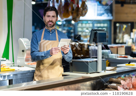 Professional shopkeeper in apron using tablet at delicatessen counter of modern grocery store. Warm smile conveys friendly service. Background shows variety of products 118190628