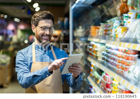 Confident grocery store worker uses digital tablet while standing in supermarket aisle. Surrounded by various products, he engages with technology to manage inventory. 118190645