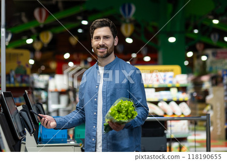 Young man using phone at self-checkout kiosk in grocery store. Person holding lettuce while smiling, engaging in digital payment process. Concept of modern technology, convenience Young man using phone at self-checkout kiosk in grocery store. Person holding lettuce while smiling, engaging in digital payment process. Concept of modern technology, convenience 118190655