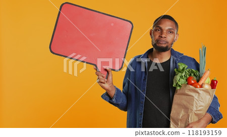 Young man showing a speech bubble empty cardboard sign to create an ad for sustainable lifestyle healthy eating. Guy doing a promotional advertisement for bio ripe fruits and veggies. Camera B. 118193159