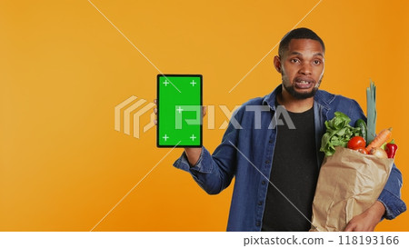Vegan adult holding a tablet with greenscreen layout in studio, carrying paper bag full of ethically sourced chemicals free produce. Guy advocating for healthy eating with mockup screen. Camera B. 118193166