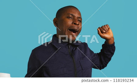 Headshot portrait of security agent drinking coffee to stop yawning while securing assigned post, studio background. Close up shot of guardian consuming caffeinated beverage, camera B 118193234