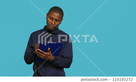 Security guard holding clipboard, taking notes, receiving orders during protection job shift. Portrait of bodyguard using pen to write informations received, studio background, camera A Security guard holding clipboard, taking notes, receiving orders during protection job shift. Portrait of bodyguard using pen to write informations received, studio background, camera A 118193272
