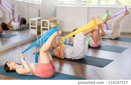 Sports girl with women and men of different ages doing stretch exercises for legs with resistance bands while lying on mats in gym Sports girl with women and men of different ages doing stretch exercises for legs with resistance bands while lying on mats in gym 118197317