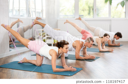 Experienced sporty young woman on hands and knees doing stretch exercises for legs with resistance bands on mats during training with group people in sports studio Experienced sporty young woman on hands and knees doing stretch exercises for legs with resistance bands on mats during training with group people in sports studio 118197417