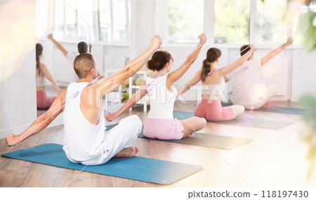 Back view of energetic active young man sitting on gymnastics mat exercising with pilates elastic resistance band with group of athletic people in fitness studio 118197430