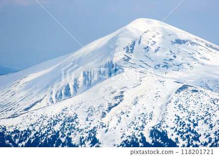 Snow-covered mountain peak under clear blue sky, with patches of snow on slopes and scattered trees. Serene winter landscape contrasts sharply with the dark, forested areas below. Hoverla, Ukraine 118201721