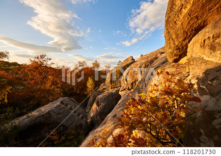 Golden hues of setting sun illuminate rocky formations and autumn foliage, casting warm glow on scene. Vibrant oranges and reds of trees below. Dovbush Rocks, Carpathian mountains, Ukraine, Europe. Golden hues of setting sun illuminate rocky formations and autumn foliage, casting warm glow on scene. Vibrant oranges and reds of trees below. Dovbush Rocks, Carpathian mountains, Ukraine, Europe. 118201730