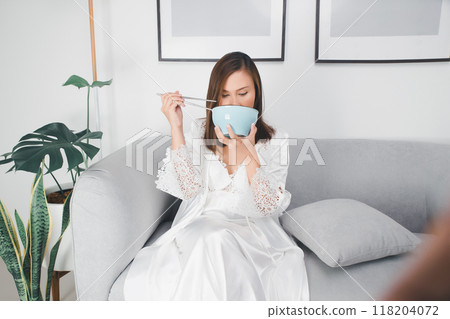 At midnight, an Asian woman, hungry and tired, sits on her couch eating food from a bowl. 118204072