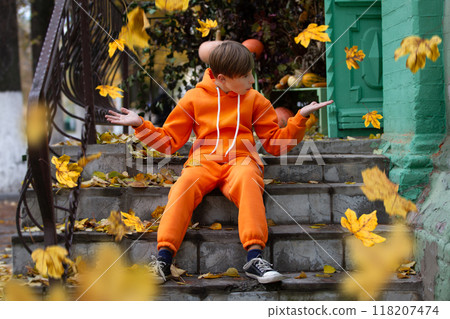 A boy in orange clothes sits on the steps and looks at the yellow flying autumn leaves. Fall has come. A boy in orange clothes sits on the steps and looks at the yellow flying autumn leaves. Fall has come. 118207474