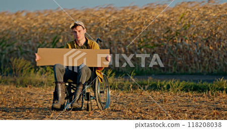 A young man in a wheelchair holds a blank cardboard sign in front of a cornfield. A young man in a wheelchair holds a blank cardboard sign in front of a cornfield. 118208038